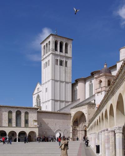 The image shows the white stone facade of the Basilica of San Francesco in Assisi, Italy, with its distinctive bell tower and rose window, alongside a modern bronze equestrian statue on a grassy lawn.