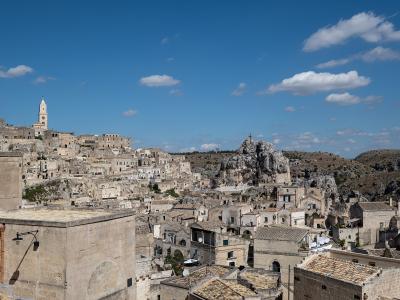 A panoramic view of the Sassi di Matera in Italy, showing ancient cave dwellings and stone buildings carved into a limestone ravine under a blue sky with scattered clouds; a prominent church bell tower is visible on the left, and a rocky outcrop w