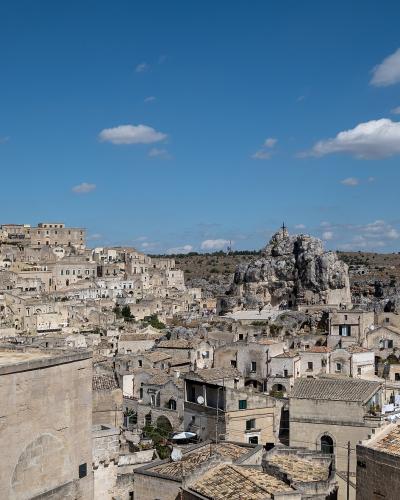 A panoramic view of the Sassi di Matera in Italy, showing ancient cave dwellings and stone buildings carved into a limestone ravine under a blue sky with scattered clouds; a prominent church bell tower is visible on the left, and a rocky outcrop w
