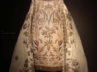 A religious vestment (dalmatic and mantellina) with embroidered gold floral patterns and a large "M" monogram, displayed on a mannequin wearing silver crowns, with museum label text visible on the left reading "62. Manifattura fiorentina" and back