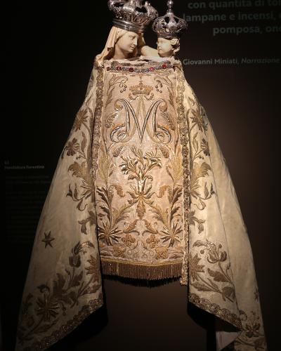A religious vestment (dalmatic and mantellina) with embroidered gold floral patterns and a large "M" monogram, displayed on a mannequin wearing silver crowns, with museum label text visible on the left reading "62. Manifattura fiorentina" and back