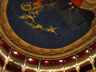The image shows the ornate interior ceiling and tiered balconies of a historic Italian opera house, featuring a large fresco with mythological figures on a blue background, gilded architectural details, red velvet drapery, and multiple levels of b