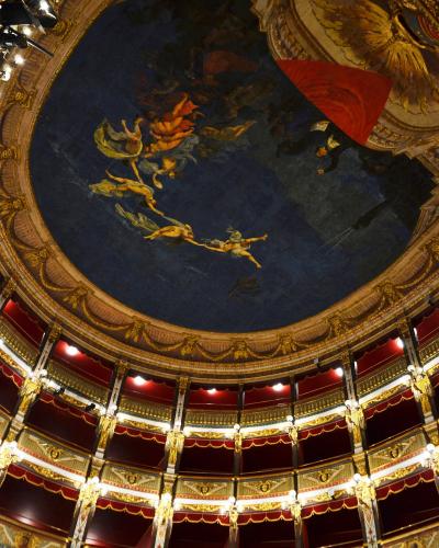 The image shows the ornate interior ceiling and tiered balconies of a historic Italian opera house, featuring a large fresco with mythological figures on a blue background, gilded architectural details, red velvet drapery, and multiple levels of b