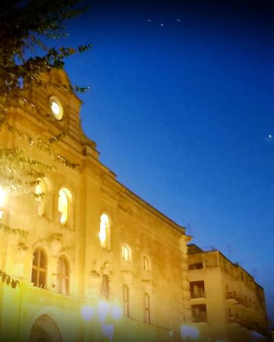 A large, illuminated stone building with a clock tower and arched windows at night, partially obscured by trees in the foreground.
