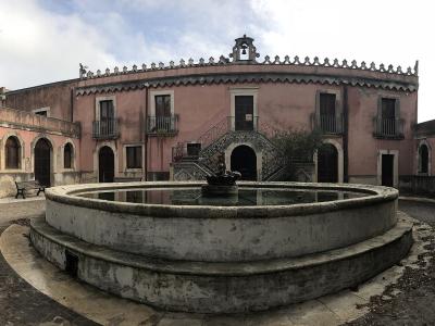 A panoramic view of a pinkish-hued historic building with a central staircase, arched loggias, and a large circular stone fountain in the foreground courtyard.