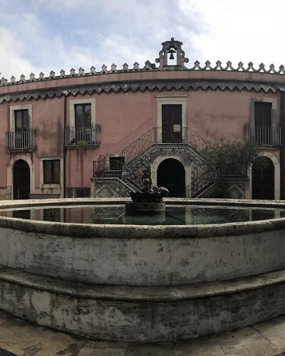 A panoramic view of a pinkish-hued historic building with a central staircase, arched loggias, and a large circular stone fountain in the foreground courtyard.
