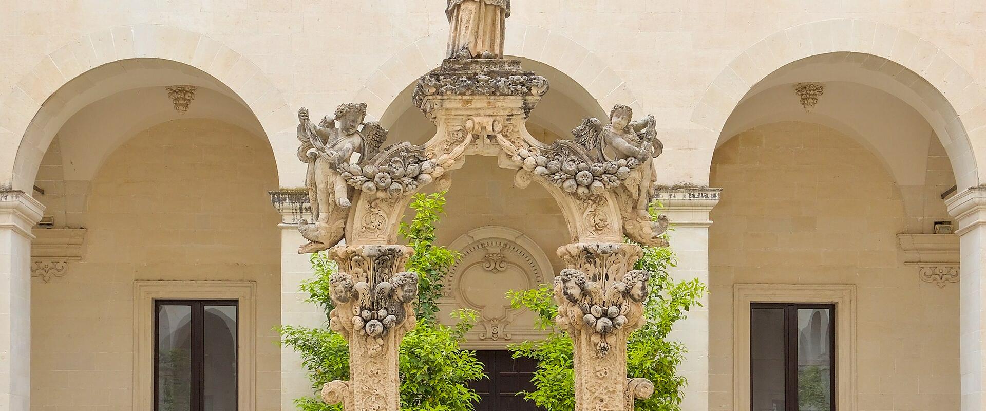 The image shows an ornate stone fountain with a statue on top, situated in a courtyard of a light-colored stone building with arched colonnades and Baroque-style architectural details.