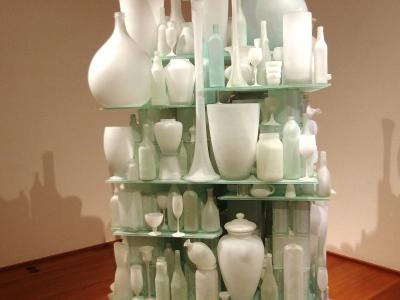 A tall, multi-tiered glass shelving unit displaying a dense collection of white and frosted glass bottles, vases, and goblets in an indoor gallery setting with wooden floors and beige walls.