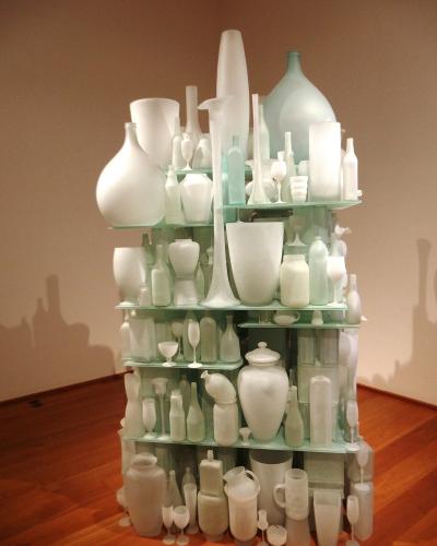 A tall, multi-tiered glass shelving unit displaying a dense collection of white and frosted glass bottles, vases, and goblets in an indoor gallery setting with wooden floors and beige walls.