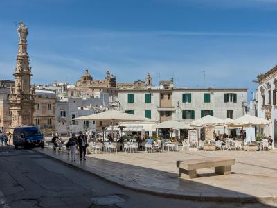 The image shows a sunlit Italian piazza featuring a prominent stone column topped with a statue on the left, outdoor café seating under umbrellas in the center, and whitewashed buildings with green shutters and a distant hilltop fortress in the ba