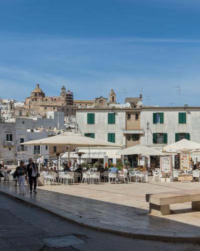 The image shows a sunlit Italian piazza featuring a prominent stone column topped with a statue on the left, outdoor café seating under umbrellas in the center, and whitewashed buildings with green shutters and a distant hilltop fortress in the ba