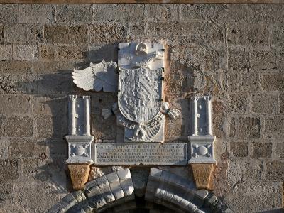 A close-up of a stone coat of arms mounted on a rough-hewn masonry wall, featuring a double-headed eagle, a shield, and an inscription referencing "CAROLO V IMPERATORE" (Charles V, Holy Roman Emperor).