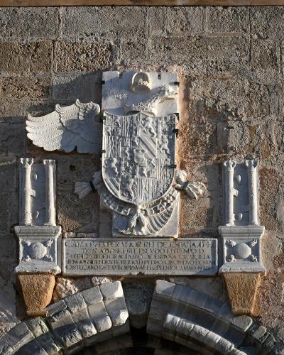 A close-up of a stone coat of arms mounted on a rough-hewn masonry wall, featuring a double-headed eagle, a shield, and an inscription referencing "CAROLO V IMPERATORE" (Charles V, Holy Roman Emperor).