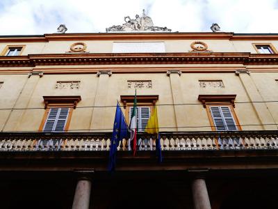 The image shows the neoclassical facade of a theater building with a central balcony, three flags (EU, Italian, and yellow), and a sculptural group on the roofline.
