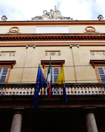The image shows the neoclassical facade of a theater building with a central balcony, three flags (EU, Italian, and yellow), and a sculptural group on the roofline.