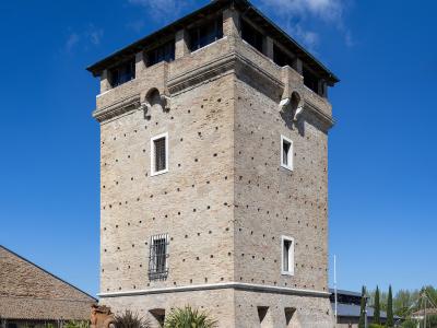 A tall, square brick tower with a wooden roof structure, set against a blue sky, with a paved plaza and some greenery at its base.